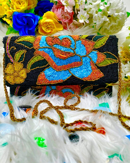 A clutch purse with a floral pattern, predominantly black with areas of blue, green, and orange, placed on a white furry surface with a selection of flowers in the background.